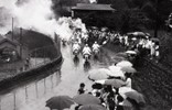 昭和39年の東京オリンピックの聖火リレー。奥多摩街道を福生方面に向かって走るランナーに声援を送る人々が沿道に集まっています。多くの人が傘を差していますが、差していない人もいます。小雨だったのでしょうか。羽村町（当時）のランナー達が走ったのは福生町境までで、約20分かけて町内を駆け抜けました。
